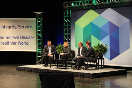 A stage with four people sitting on it. The stage is decorated with a large screen on the right side and a banner on the left side that reads "Integrity Service and Relieve Disease Healthier World." The people on the stage are dressed in formal business attire and appear to be engaged in a discussion. There are two potted plants on either side of the stage. The background is dark suggesting that the event is taking place at night.