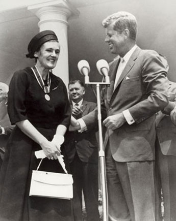 A black and white photograph of a man and a woman standing in front of a podium with microphones. The woman is on the left side of the image wearing a black dress and a hat and is holding a white handbag. She is smiling and appears to be giving a speech or presentation to the man on the right side. The man is wearing a suit and tie and is looking at the woman with a smile on his face. Behind them there are several other people standing and clapping and a column can be seen in the background. The image appears to have been taken at a political rally or event.