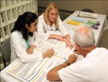 Three people two women and an elderly man sitting at a table and having a discussion. They are all wearing white lab coats and appear to be in a medical office or clinic setting. The table is covered with a white tablecloth and there are several papers and brochures scattered across it. The woman on the left is a young woman with long dark hair the woman in the middle is a blonde woman with shoulder-length hair and the man on the right is an older man with white hair. All three people are looking at the papers and seem to be engaged in the conversation.