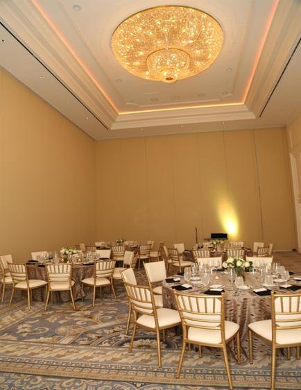 A large ballroom with a high ceiling and a chandelier hanging from it. The room is set up for a formal event with round tables and chairs arranged in a semi-circle. The tables are covered with white tablecloths and have white flower arrangements on top. The floor is covered with a blue and beige patterned carpet. The walls are painted in a light beige color and there is a large window on the right side of the room letting in natural light. The overall atmosphere is elegant and sophisticated.