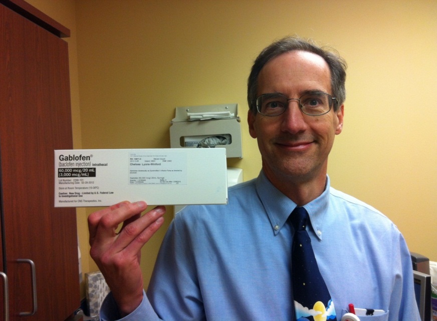 A middle-aged man in a blue shirt and tie holding up a white card with the name "Gablofen" written on it. He is standing in a room with a yellow wall and a wooden cabinet in the background. The man is smiling and looking at the camera. He appears to be in his late 40s or early 50s and has short dark hair and glasses. On the right side of the image there is a desk with a computer monitor and other office supplies.