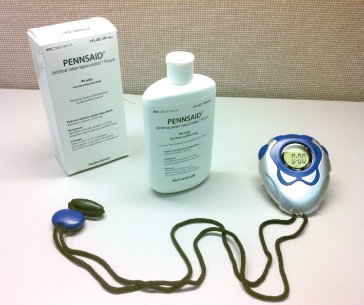 A white box with the word "PENNSAID" written on it. Next to the box there is a white bottle with a white cap and a blue label. The bottle has a label that reads "PENNSAID" in black letters. On the right side of the image there are two small blue objects one of which appears to be a stopwatch and the other of which is connected to a green cord. The stopwatch has a digital display that shows the time. The background is a beige-colored surface.