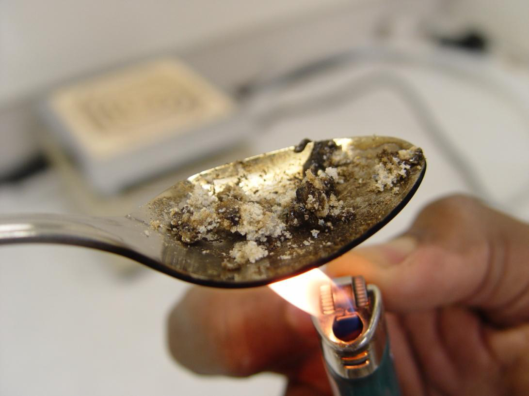 A silver spoon with a small amount of white powder on it likely heroin or another drug. A person is holding a lit lighter under the spoon and the powder seems to be darkening in color due to the heat. The background is blurred but it appears to be light in color and possible a kitchen or laboratory setting.