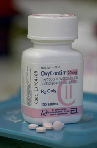 A white prescription bottle with a pink label. The label reads "OxyContin 20 mg (oxycodone hydrochloride controlled-release tablets) Rx Only". The bottle has a white cap and is placed on a blue tray. There are a few white pills scattered around the bottle. The background is blurred but it appears to be a medical setting.