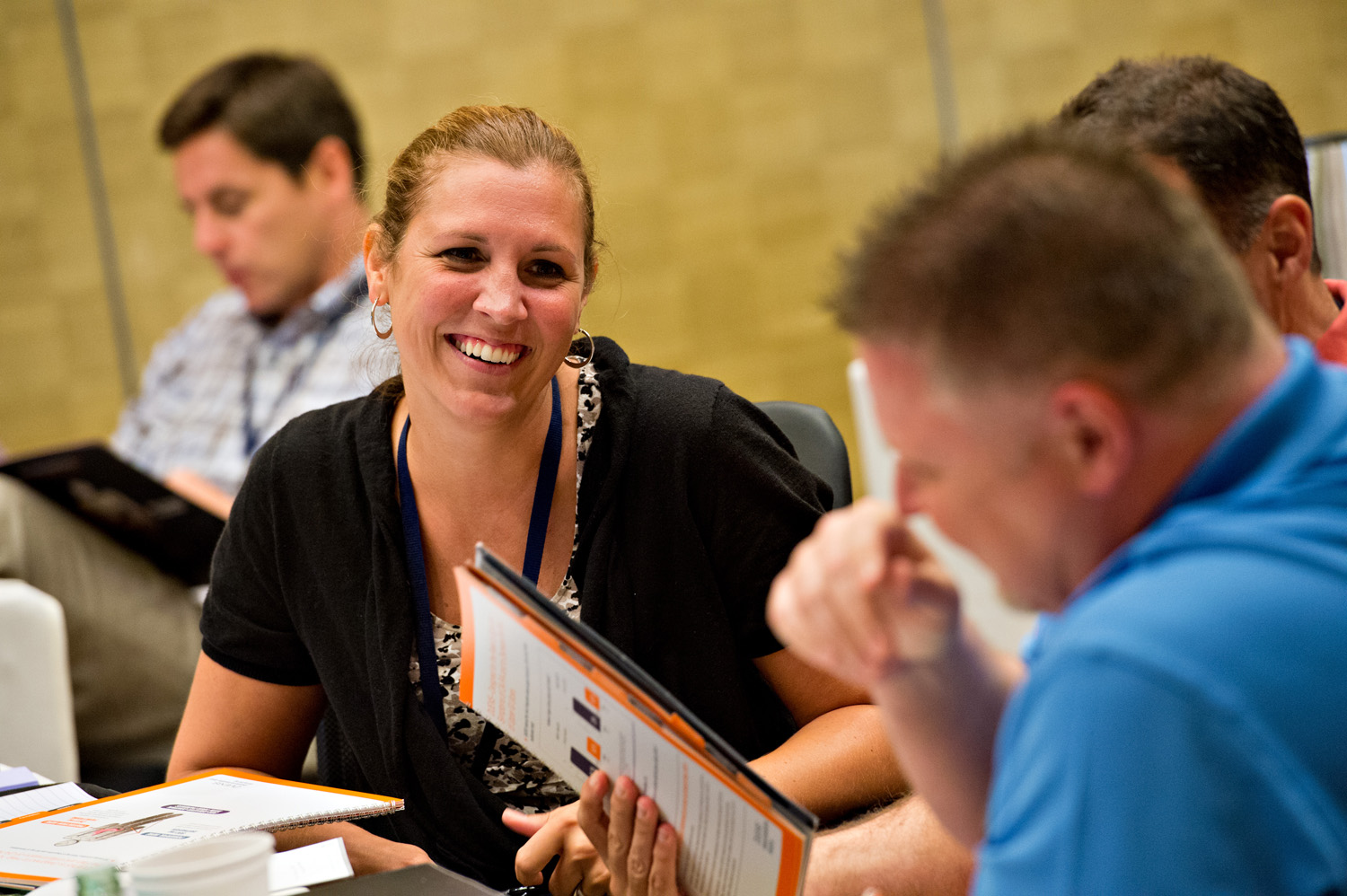 A group of people sitting at a table in a conference room. There are three people in the image two men and one woman. The woman is in the center of the image and is smiling at the camera. She is wearing a black top and has a lanyard around her neck. The man on the right is holding a folder and appears to be deep in thought. The other two men are sitting on either side of her also smiling. They are also holding folders and appear to be engaged in a discussion. The table is covered with a white tablecloth and there are papers and a cup of coffee on it. The background is blurred but it seems like the room is well-lit with natural light coming in from the windows.