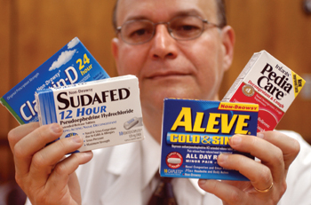 A middle-aged man wearing a white lab coat and glasses holding three boxes of medicine in his hands. The first box on the left is labeled "Sudafed 12 Hour" and has a blue and white label with the words "Pedia Care" written on it. The second box is titled "Aleve Cold & Sin" and is labeled as "All Day Pain Relief". The third box is labeled as "Pediatric Care". The man is smiling and looking at the camera. The background is blurred but it appears to be a room with a wooden paneled wall.