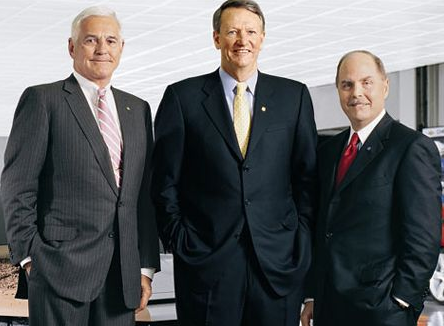 Three men standing in an office setting. They are all dressed in formal attire wearing suits and ties. All three men are smiling and appear to be posing for the photo. In the background there is a desk with a computer monitor and other office supplies. The overall mood of the image is professional.