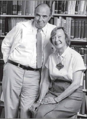 A black and white photograph of an elderly couple posing for a photo in front of a bookshelf. The man is on the left side of the image wearing a white shirt and tie and the woman is sitting on the right side. They are both smiling and looking at the camera. The woman has short light-colored hair and is wearing a necklace with a pendant. The bookshelves behind them are filled with books of various sizes and colors. The image appears to be taken in a library or study setting.