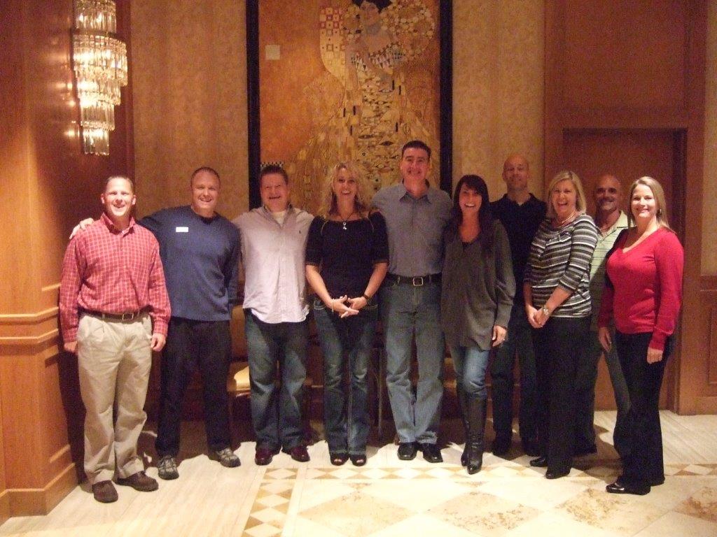 A group of nine people standing in a large room with a high ceiling and a chandelier hanging from the ceiling. The room appears to be a lobby or reception area with a marble floor and wooden walls. There is a large painting hanging on the wall in the background. The people in the group are of different ages and ethnicities and they are all smiling and posing for the photo. They are all dressed in casual clothing and appear to be happy and relaxed.