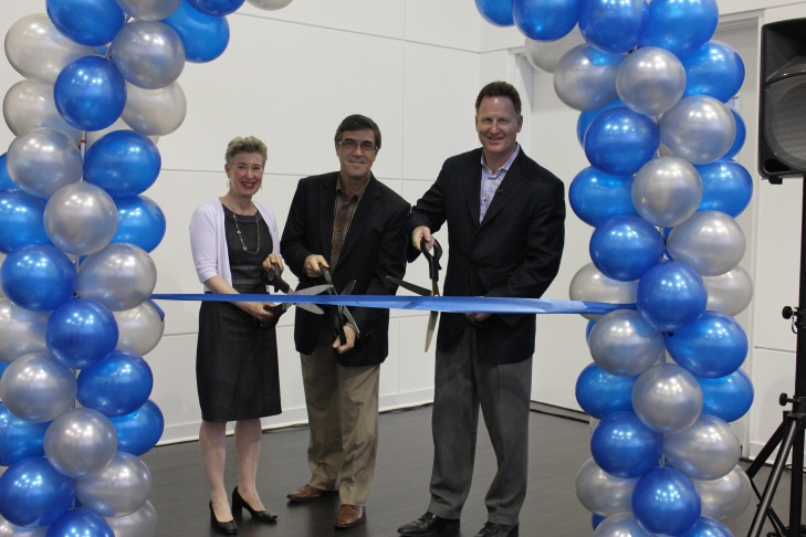 Three people two men and a woman standing in front of a large arch made of blue and silver balloons. They are holding a pair of scissors and are cutting a blue ribbon that is tied in a bow. The arch is set up in a room with a white wall and a speaker on a tripod stand in the background. The people are smiling and appear to be celebrating a ribbon cutting ceremony.