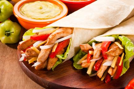 Two wraps on a wooden cutting board. The wraps are filled with grilled chicken red and yellow bell peppers lettuce and cheese. The chicken appears to be seasoned with herbs and spices and the cheese is melted and bubbly. There are also two small bowls of dipping sauces on the cutting board one with a yellow sauce and the other with a red sauce. The cutting board is surrounded by green and red bell peppers. The background is a wooden table.