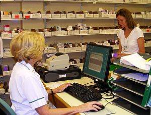 Two women in a pharmacy. The woman on the left is sitting at a desk with a computer monitor and keyboard in front of her. She is wearing a white uniform and appears to be a pharmacist or a customer service representative. The other woman is standing behind the desk and is looking at the computer screen. On the desk there is a printer and some papers. In the background there are shelves stocked with various medicines and supplies. The overall atmosphere of the image is professional and organized.