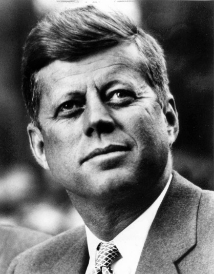 A black and white portrait of a man. He appears to be in his late 50s or early 60s with short neatly combed hair and a serious expression on his face. He is wearing a suit and tie and is looking off to the side with a slight smile on his lips. The background is blurred so the focus is on the man's face.