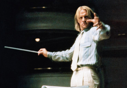 A man standing on a stage with a baton in his right hand. He is wearing a light blue shirt and beige trousers. He has long blonde hair and is looking directly at the camera with a serious expression on his face. The background is dark and blurred but it appears to be a concert hall or auditorium. The man is holding the baton with both hands and is pointing upwards with his index finger. There is a sheet of music on the stage in front of him.