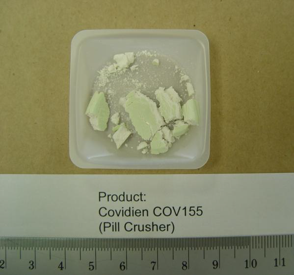 A small white plastic container with a label that reads "Product: Covidien COV155 (Pill Crusher)". Inside the container there is a pile of white powder which appears to be a type of pill crusher. The powder is finely ground and has a rough texture. The container is placed on a beige surface with a ruler next to it indicating that the product is being measured.