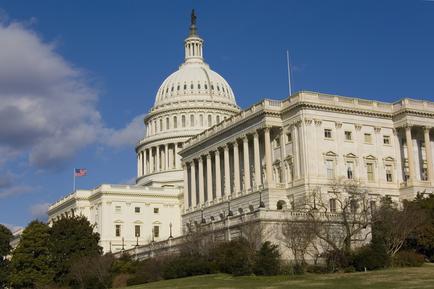 A photograph of the United States Capitol building in Washington DC. The building is a large neoclassical structure with a dome-shaped dome on top and a statue of a man on a pedestal on the right side. The dome is surrounded by columns and arches and there is a flagpole with the American flag flying on the left side of the image. The sky is blue with a few clouds and the building is located on a hill with trees and shrubs in the foreground. The image is taken from a distance looking up at the building.