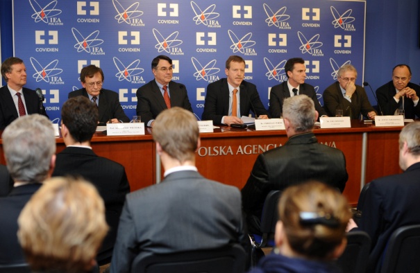 A group of nine men sitting at a long table in front of a blue backdrop with the logo of the Helsinki Agency. The men are dressed in formal business attire and appear to be engaged in a discussion. The table is covered with a red tablecloth and there are nameplates and microphones on it. The audience is seated in rows of chairs facing the table. The image appears to be taken at a conference or meeting.