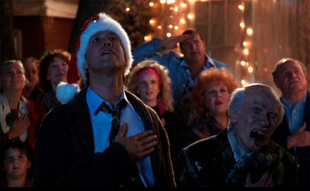 A group of people standing in front of a Christmas tree decorated with lights. In the center of the image there is a man wearing a red Santa hat and a black suit. He is standing with his arms crossed and appears to be singing into a microphone. To his right there are two older men one with white hair and the other with gray hair. Behind them a crowd of people can be seen clapping and cheering. The background is dark suggesting that the photo was taken at night.