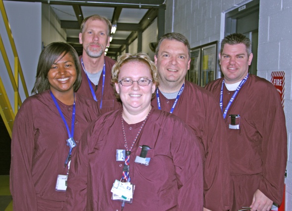 A group of five people standing in a hallway. They are all wearing maroon scrubs and have blue lanyards around their necks. The person in the front is a woman with blonde hair and glasses and she is smiling at the camera. Behind them there are two men and two women all of whom are also smiling. On the right side of the image there is a fire extinguisher on the wall. The hallway appears to be empty with no other people or objects visible in the background.