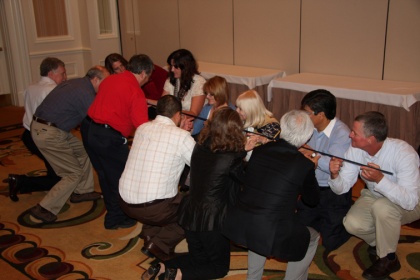 A group of people gathered in a room with a carpeted floor and a white tablecloth in the background. There are nine people in the image six men and three women all of whom appear to be engaged in a physical altercation. The men are kneeling on the floor while the women are standing around them some of them are holding swords. They are all looking at each other and seem to be in the middle of a fight. The overall mood of the image is tense and confrontational.