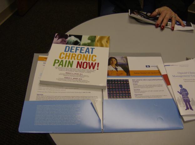 A table with a few items on it. On the left side of the table there is a book titled "Defeat Chronic Pain Now!" with an image of a woman's face on the cover. Next to the book there are two pamphlets with text and images. The pamphlets appear to be related to the topic of chronic pain. The table is covered with a white tablecloth and there are a few papers scattered around it. A person's hand is visible in the top right corner of the image holding a pen and writing on one of the pamphlets.