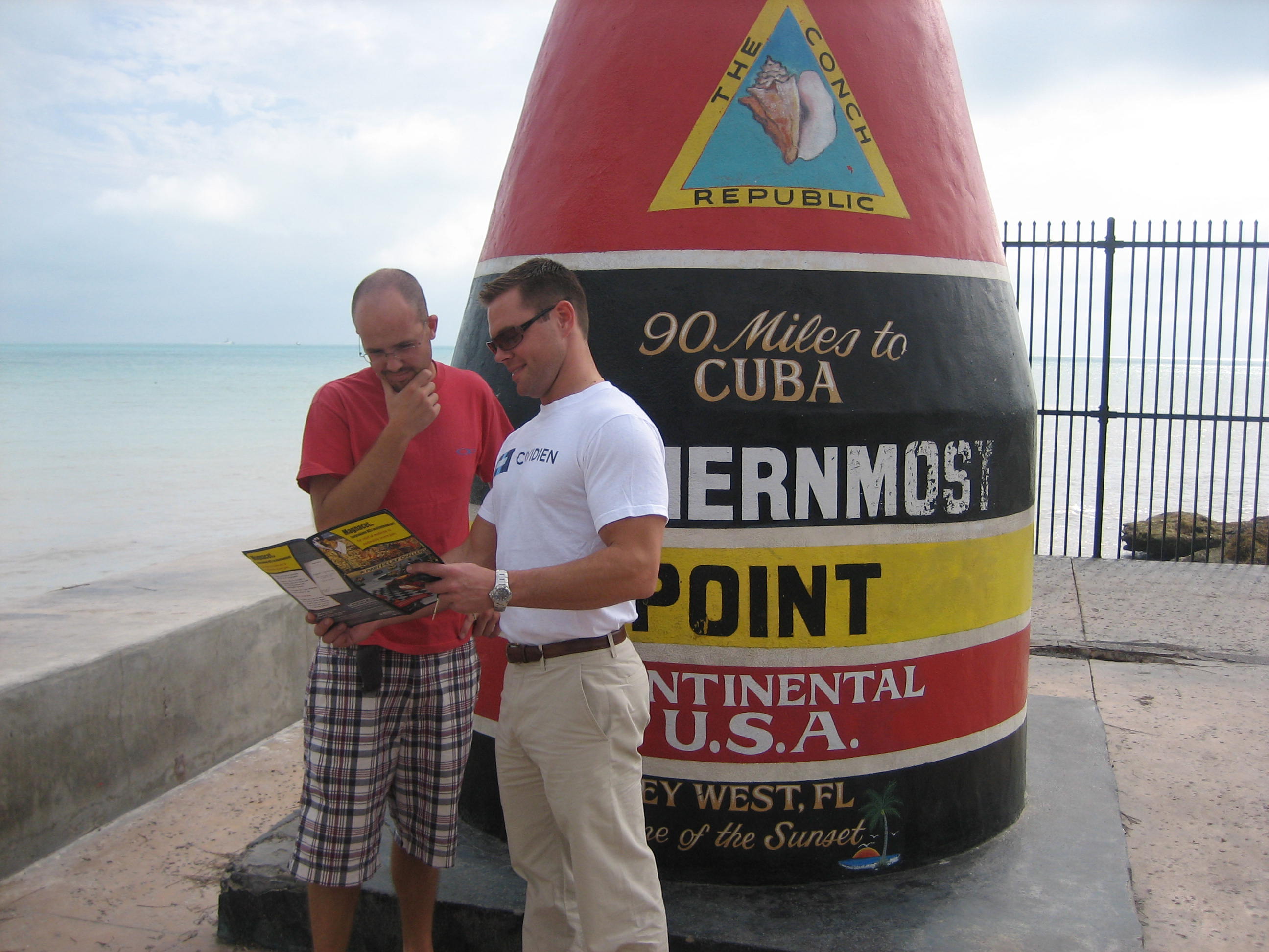 Two men standing in front of a large red and yellow sign that reads "90 Miles to Cuba" and "International Point Continental USA". The sign is located on a concrete wall near the ocean with a black fence in the background. The man on the left is wearing a red t-shirt and plaid shorts and is holding a map in his hand. He is looking at the map with a smile on his face while the other man is standing next to him wearing a white shirt and khaki pants. Both men appear to be looking at it intently. The sky is blue and the ocean can be seen in the distance.