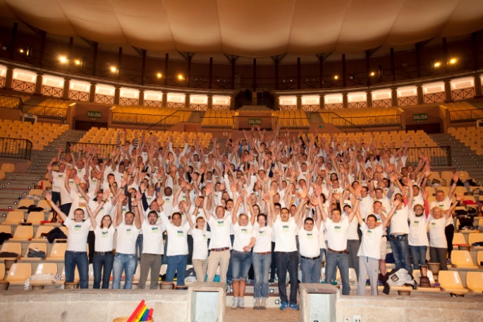 A large group of people standing on a stage in a large auditorium with rows of empty seats. They are all wearing white t-shirts with the logo of a company or organization on them. The people are all raising their hands in the air and appear to be cheering or celebrating. The stage is made of concrete and there is a podium in front of the group. The auditorium has a high ceiling with a curved design and is lit up with bright lights.