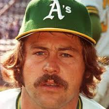 A close-up portrait of a man wearing a green baseball cap with the letter "A's" on it. He has long curly hair and a mustache. He is wearing a white jersey with green stripes on the sleeves. The background is blurred but it appears to be a baseball stadium with spectators in the stands. The man is looking directly at the camera with a serious expression on his face.