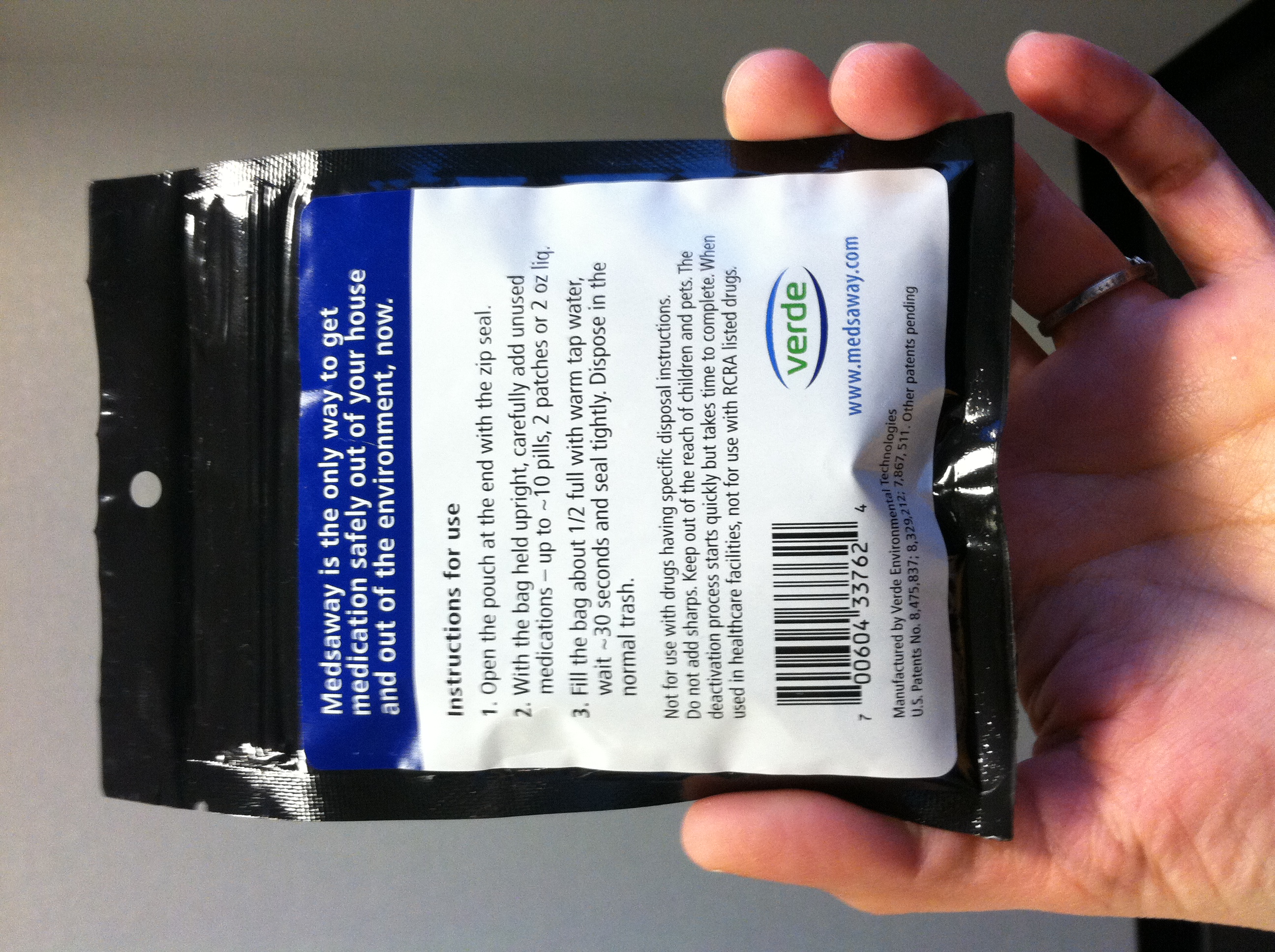 A hand holding a black plastic packet with a blue and white label on it. The label has the brand name "Verde" written in bold white letters at the top followed by the product name and a barcode. Below the label there is a description of the product which states that it is made with natural ingredients and is suitable for use in a variety of applications. The packet appears to be sealed.