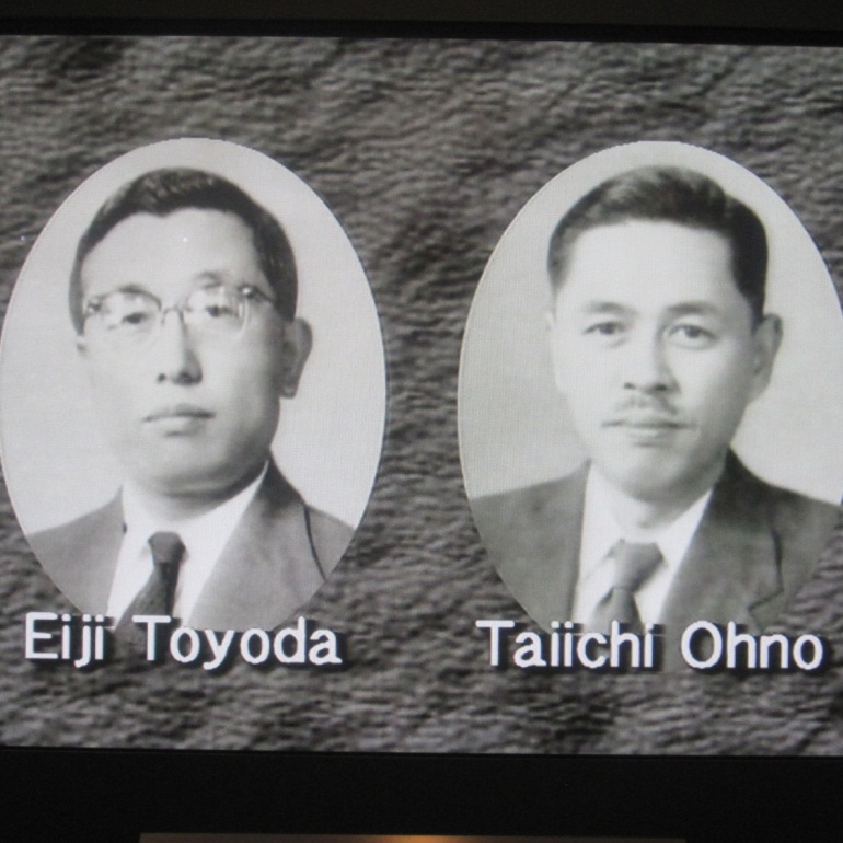 The image has two black and white photographs each a professional headshot. They are of two men Eiji Toyoda and Taiichi Ohno side by side. They are both wearing suits and ties and are facing the camera. The photograph is circular in shape and has a white background. The man on the left is wearing glasses and the man on the right has a mustache. 