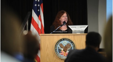 A woman standing behind a wooden podium with the seal of the United States of America on it. She is speaking into a microphone and appears to be giving a speech. The background is dark and there is an American flag on the left side of the image. The woman has long red hair and is wearing a black blazer. There are people sitting in front of the podium attentively listening to her speech.