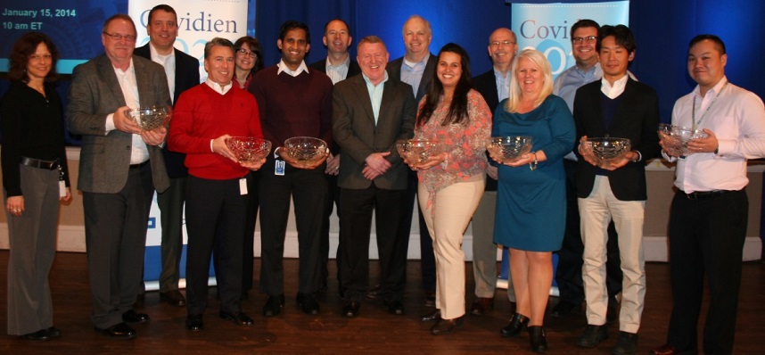 A group of people standing on a stage with a blue curtain in the background. There are nine people in the group nine men and one woman all of whom are holding silver bowls. They are all smiling and appear to be posing for a photo. The man in the center is wearing a red sweater and is holding a trophy while the others are holding smaller bowls. On the left side of the image there is a banner that reads "Covidien" and on the right side there are two banners that read "January 15 2014". The group appears to be at an awards ceremony or recognition event.