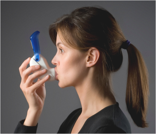 A young woman with shoulder-length brown hair tied in a ponytail. She is wearing a black top and is holding a blue and white inhaler in her hand. The background is a plain grey color. The woman appears to be in her late twenties or early thirties.