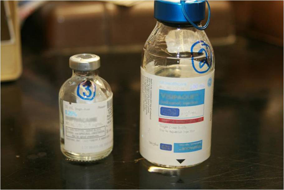 Two small bottles of water on a black countertop. The bottle on the left is a clear glass vial with a silver cap and a blue label. The label has a blue and white logo of a blue circle with the letter "S" in the center. On the right is a transparent glass bottle with a blue cap. Both bottles have a white label with blue and red text. The background is blurred but it appears to be a kitchen countertop with a sink and other kitchen appliances visible.