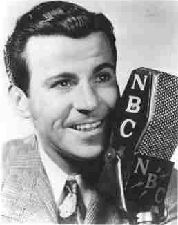 A black and white photograph of Dennis Day an actor holding a microphone. He is wearing a suit and tie and has a big smile on his face. He appears to be in his late twenties or early thirties. The microphone has the letters "NBC" on it indicating that it is a news broadcast microphone. The background is plain and white making the man and the microphone the focal point of the image.