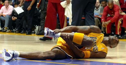 A basketball player from the Los Angeles Lakers lying on the court with his foot shin and ankle in his hands. He is wearing a yellow jersey with the team's logo on it and yellow shorts. He appears to be in pain. In the background there are spectators in the stands and a referee on the sidelines. The image appears to have been taken during a game.