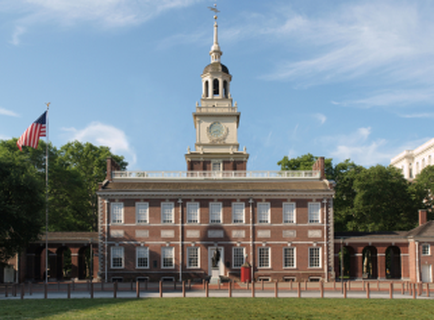 A large brick building with a clock tower on top. The building has a dome with a cross on top and a clock face in the center. The clock tower is surrounded by a white railing and there is an American flag flying on the left side of the building. The sky is blue and there are trees in the background. The ground is covered in green grass.