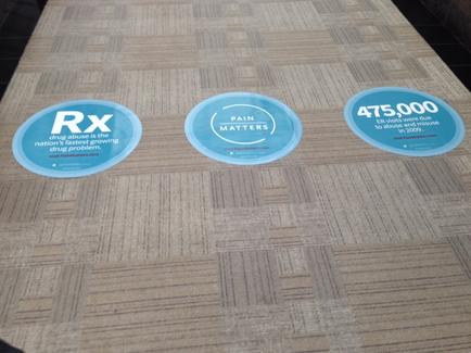 Three circular stickers on a beige carpeted floor. The stickers are blue in color and have the text "RX" "Pain Matters" and "475000" written on them. The text on the stickers is in white and is in a bold font. The background of the image is blurred but it appears to be an indoor space with a black tiled floor.