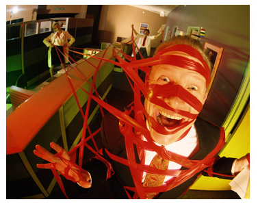 A man in an office cubicle with his mouth open wide as if he is screaming or shouting. He is wearing a suit and tie and has a red bandage wrapped around his head and neck. The bandage appears to be made up of multiple red ribbons or streamers creating a web-like effect. The man's face is covered in red tape and his eyes are wide open and his mouth is slightly open showing his teeth. In the background there are other people in the cubicle some of whom are also covered in the same red tape. The overall mood of the image is chaotic.