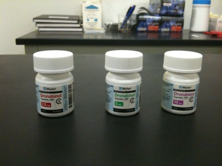 Three white plastic bottles with white caps on a black countertop. The bottles are labeled with the  name "Dronabinol". The bottles appear to be new and unused. In the background there is a desk with various items on it including a blue box a pen and a pair of scissors. The desk appears to be in a laboratory or laboratory setting.