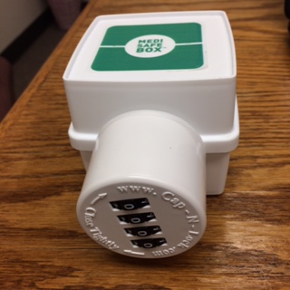 A white box with a green label on top. The label reads  "MEDI SAFE BOX". The box has a combination lock protruding from it in a cylinder shape. Around the lock the following text is in a circle: "www.Cap-N-Lock.com Close Tightly". 