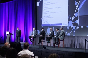 A panel discussion on a stage with a blue curtain in the background. On the left side of the stage there is a man standing at a podium and speaking into a microphone. He is wearing a suit and tie and appears to be giving a presentation. <br /><br />On the right side there are six men sitting on chairs facing the audience. They are all dressed in formal business attire and appear to be engaged in a discussion. The audience is seated in rows of chairs facing the stage.<br /><br />In the center of the image the man at the podium is speaking into the microphone and is gesturing with his hands as he speaks. Behind him the screen displays a presentation slide with a white background and blue lines and dots.<br /><br />Overall the image appears to have been taken at a conference or presentation as the audience is attentively listening to the presentation.