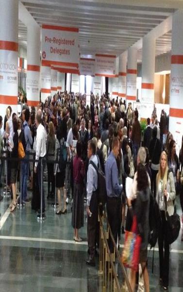 A large group of people standing in a long hallway with orange and white banners hanging from the ceiling. The banners have the words "Pre-Registered Delegates" written on them in bold black letters. The hallway appears to be an exhibition hall or convention center as there are several columns on either side of the hallway. The people in the image are of different ages genders and ethnicity's and they are all looking towards the banners. Some of them are carrying bags suggesting that they are attending an event or gathering. The overall mood of the image is busy and bustling.
