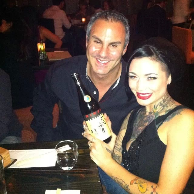 A man and a woman sitting at a table in a restaurant. The man is holding a bottle of champagne and the woman is smiling at the camera. They are both dressed in formal attire with the man wearing a black shirt and the women wearing black dresses. The table is covered with a black tablecloth and there are other people sitting at tables in the background. The atmosphere appears to be relaxed and casual.