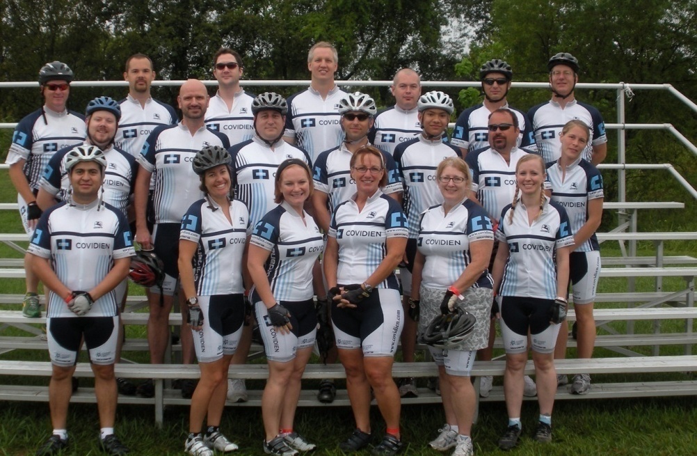 A group of people standing on a set of bleachers. They are all wearing blue and white cycling jerseys and helmets. Some of the people are holding helmets while others are standing with their hands on their hips. They appear to be posing for a group photo. The bleachers are located on a grassy area with trees in the background. The sky is overcast and the overall mood of the photo is casual and relaxed.