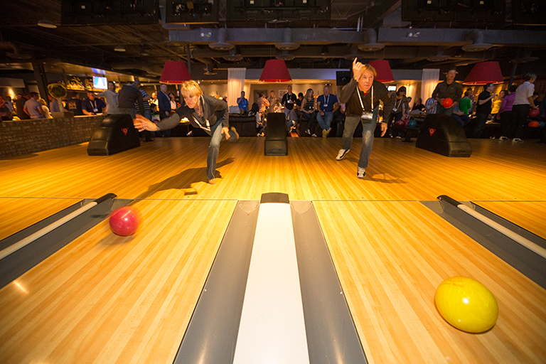 A bowling alley with wooden lanes and red and yellow bowling balls. In the center of the image there are two people a man and a woman in the middle of a bowling match. The man is in the process of throwing the ball while the woman is behind him. They are both wearing casual clothes and appear to be focused on the ball.<br /><br />In the background there is a large crowd of people watching the game. The bowling alley is dimly lit with red lamps hanging from the ceiling. There are also several bowling pins visible in the background. The image appears to have been taken at night as the lights are on and the atmosphere is lively.