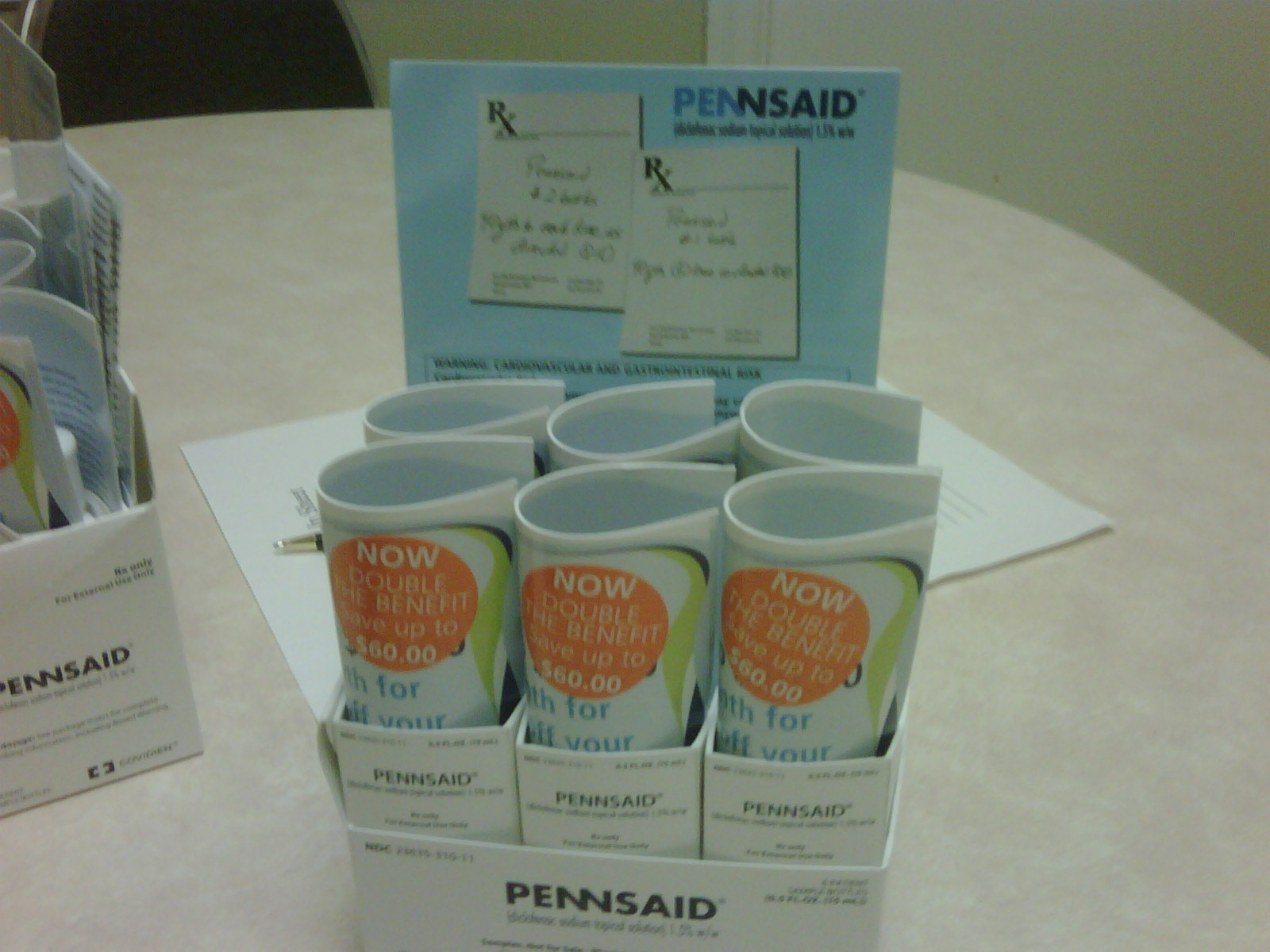 A display of Pennsaid-branded products on a table. There are six pens in a white cardboard box with the brand name "Pennsaid" written on it. The box is rectangular in shape and has a blue background with the word "Pennsaid" written in white letters. The pens are arranged in a neat row and appear to be new and unused. On the left side of the box there is a small white box with a pen in it and on the right side there are a few pens and a notepad. In the background there appears to be a desk or table with a computer monitor and other office supplies.