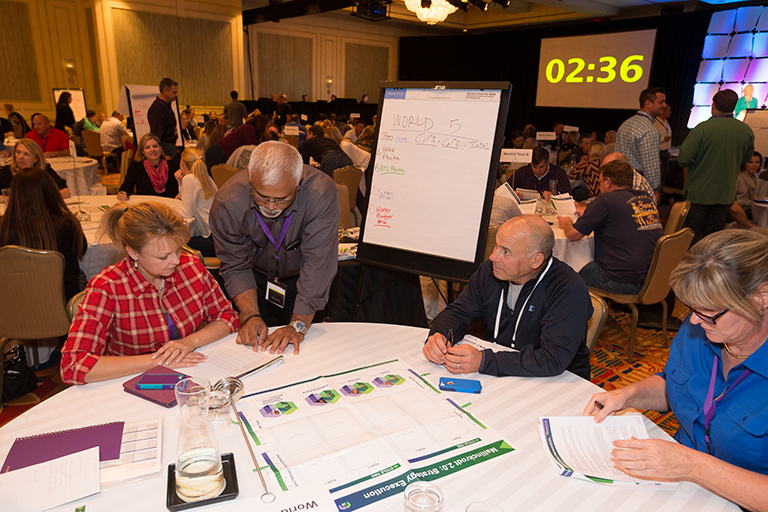 A group of people sitting at a table in a conference room. There are four people in the image three men and two women all of whom appear to be engaged in a discussion. The table is covered with a white tablecloth and there is a large whiteboard in front of them with the date "02:36" written on it. The people are looking at the whiteboard and seem to be working on a project or presentation. There is a chandelier hanging from the ceiling and a large screen in the background. The room is filled with other people some of whom are also sitting at tables and some are standing.