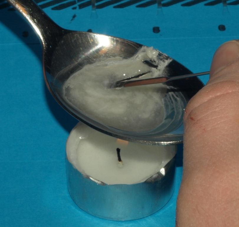 A person's hand holding a silver spoon over a small white tea candle. The spoon is filled with a white substance likely heroin or another drug. The candle is lit and the background is a blue surface. On the right side of the image a person's hand appears to be stirring the substance with what appears to be a metal needle or nail.