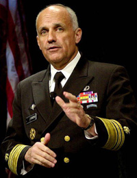 A man in a military uniform standing on a stage with an American flag in the background. He is wearing a dark green blazer with gold epaulettes and a white shirt with a black tie. He has several medals and insignia on his chest including a badge with the letter "D" on it. The man is gesturing with his hands as if he is giving a speech or making a point. He appears to be bald and has a serious expression on his face.